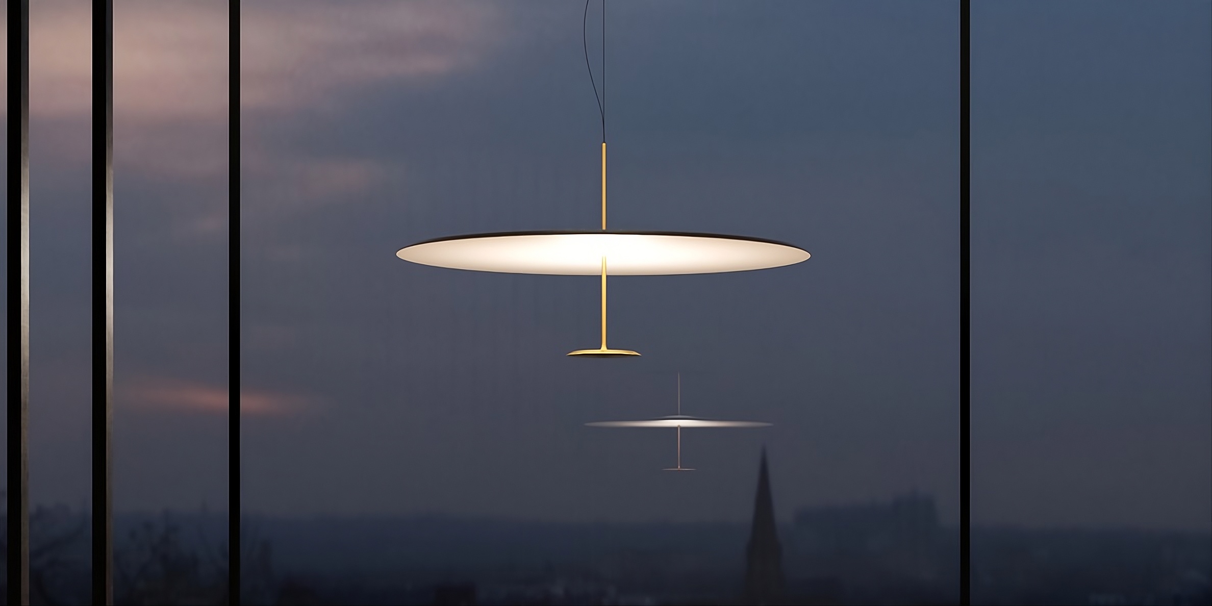 Moderne LED-Pendelleuchte mit flachem Schirm in Weiß und Gold, hängend vor Fensterfront mit Abendhimmel