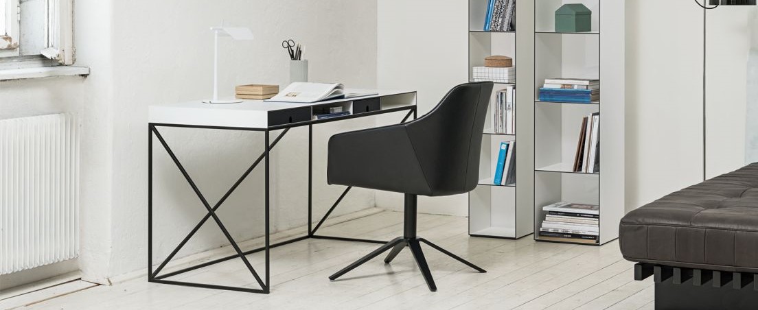 Wogg Caro Schreibtisch aus schwarzem Metall und weißer Platte in heller Raumsituation mit schwarzem Stuhl und Regal.