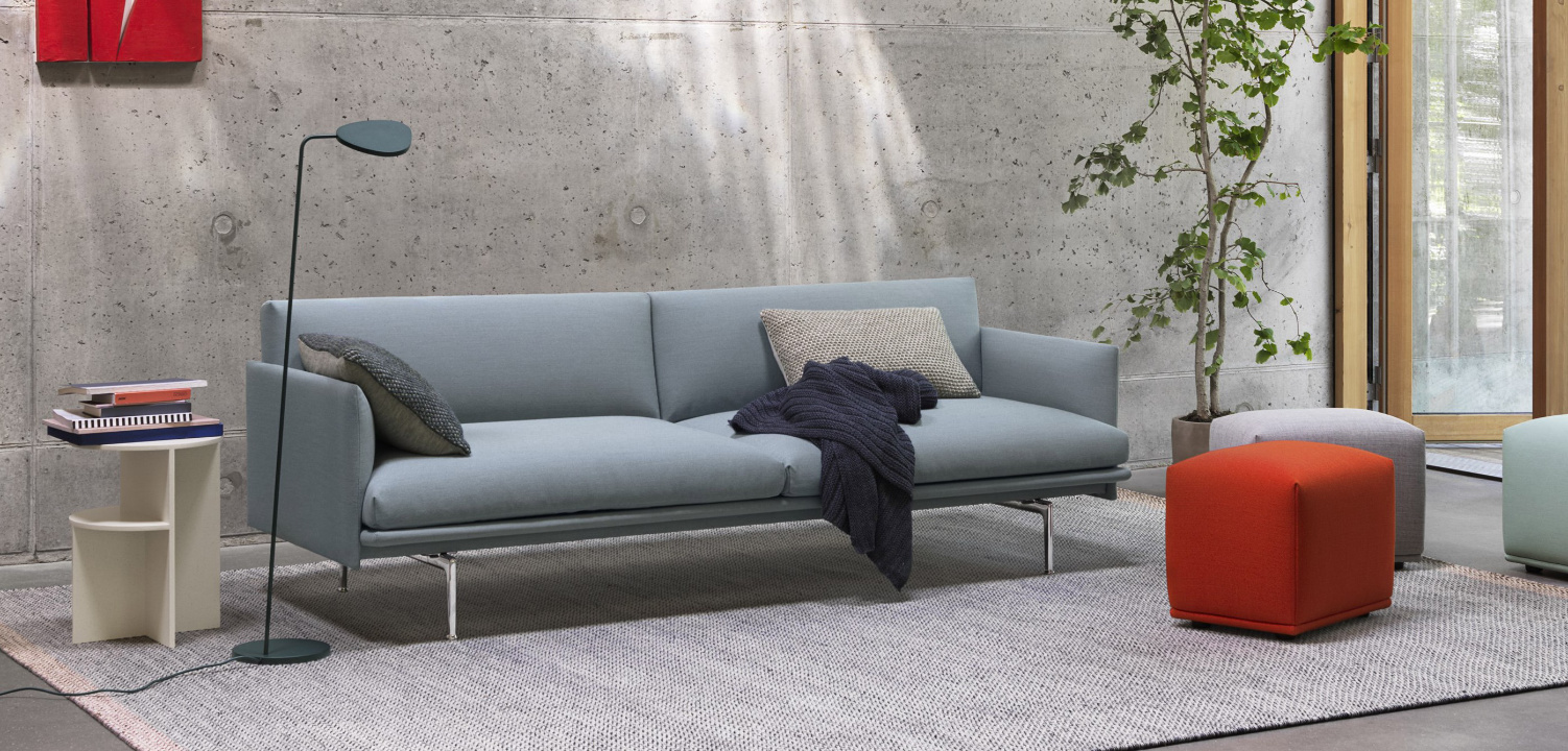 Modernes Wohnzimmer mit grauem Muuto Sofa, rotem Pouf und Betonwand in Frontalansicht.